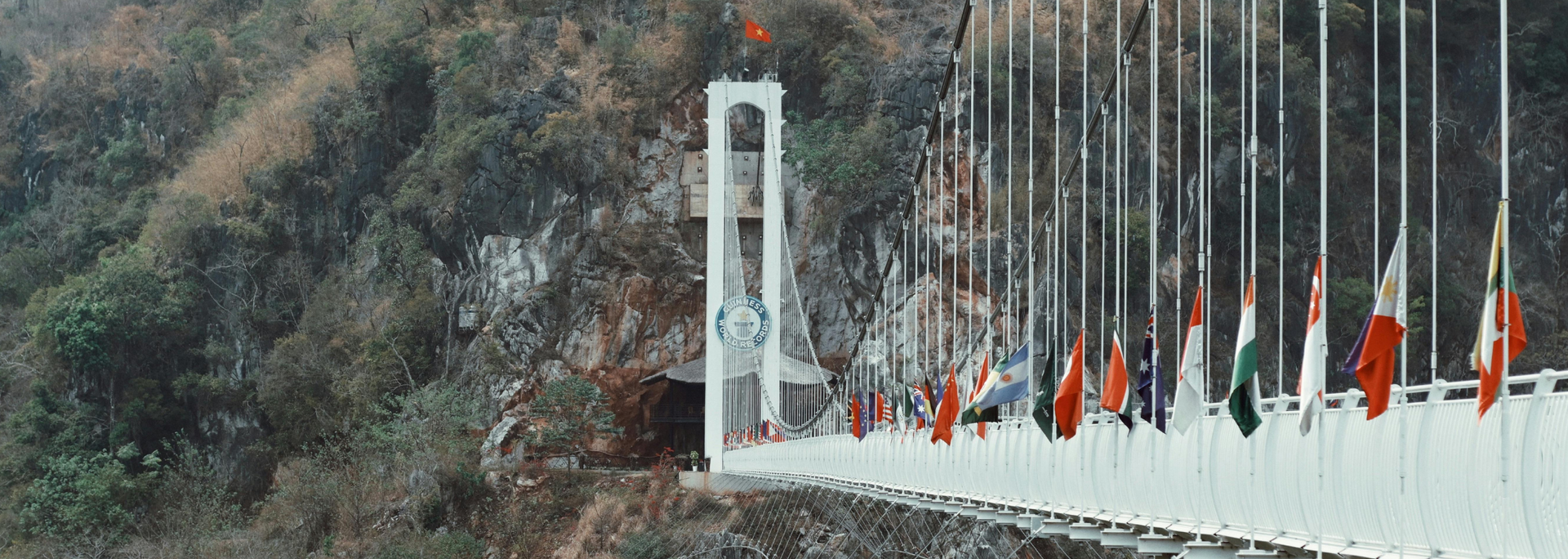 Bridge with multiple flags waving, set in a mountain landscape with a white arched structure.