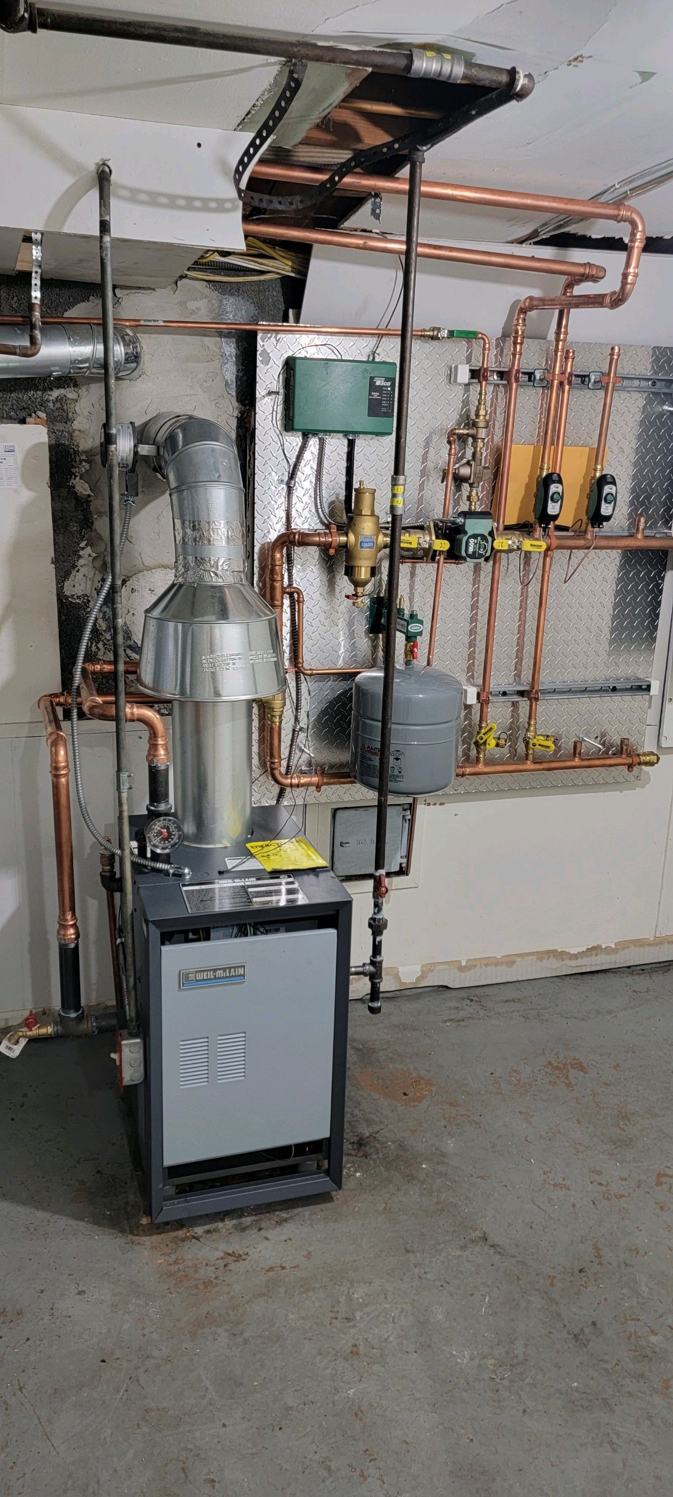 A boiler is sitting in a basement next to a wall with copper pipes.