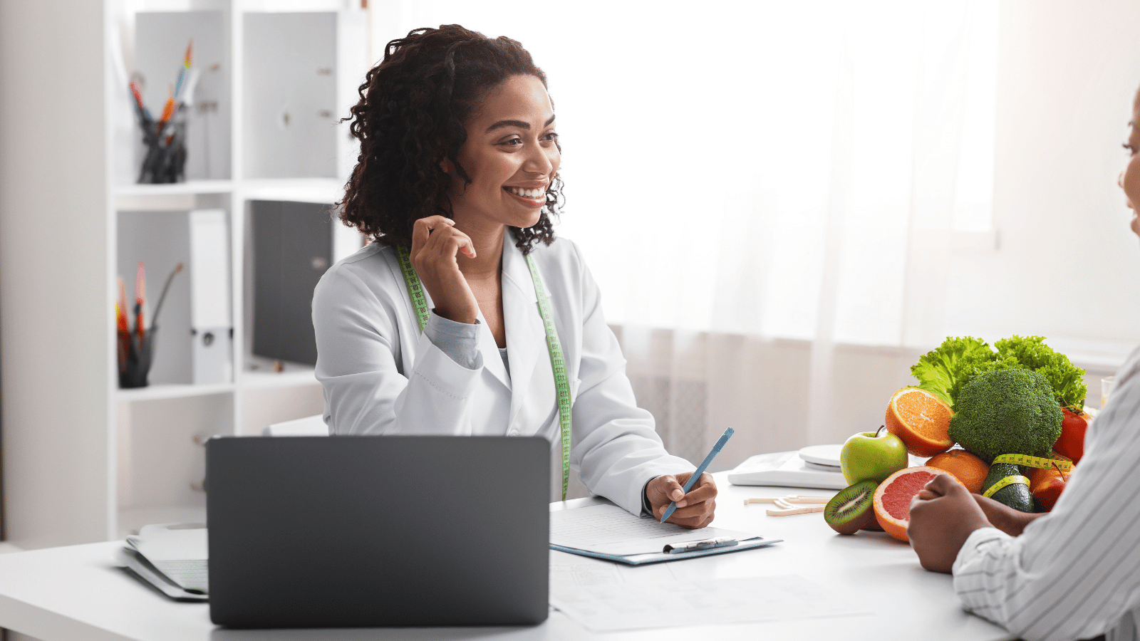 Dietitian in lab coat smiles, talking to a patient at a desk, with a laptop and fruit.