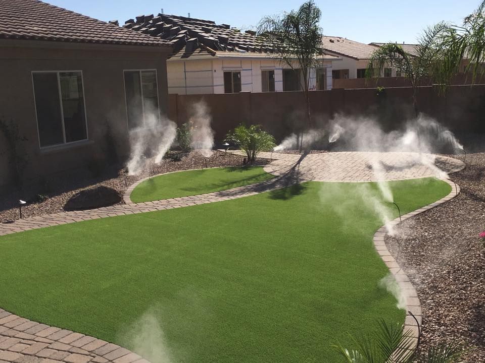 Backyard with green turf and misting spray nozzles; surrounded by brick and stone, under a blue sky.