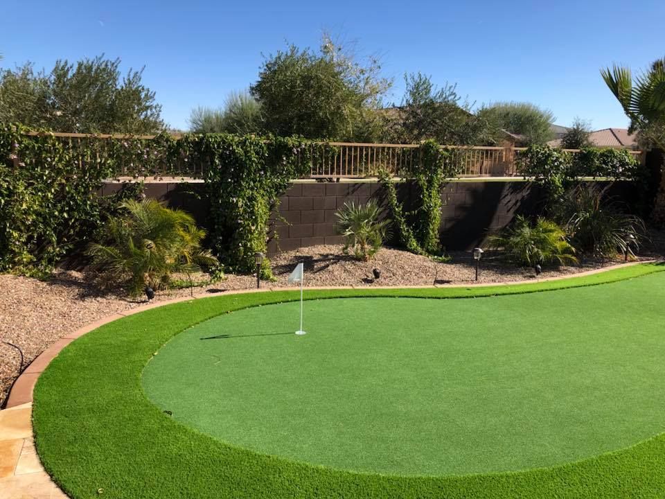Artificial turf putting green in a backyard, with surrounding landscaping and retaining wall, under a blue sky.