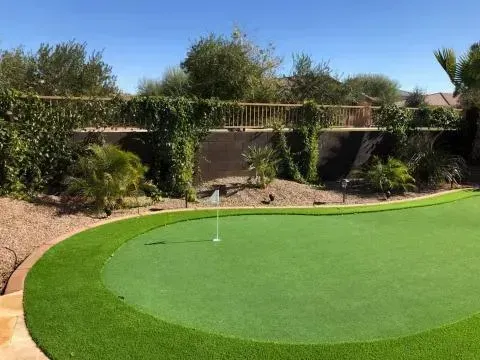 Artificial turf putting green in a backyard, with a flag and retaining wall lined with vines.