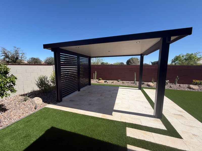 Black covered patio structure with slatted side walls over a light stone patio in a backyard with artificial grass.