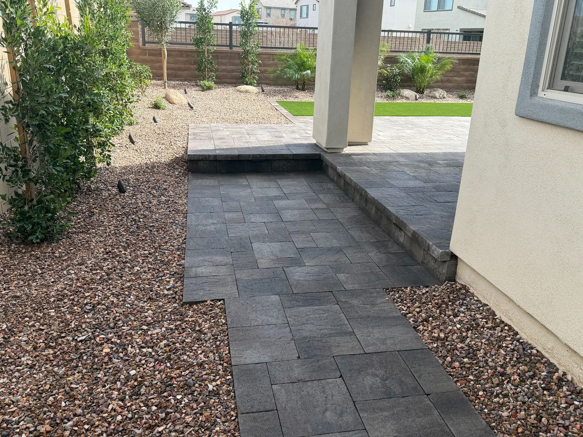 Stone paver walkway beside a beige house, bordered by gravel and shrubs in a small backyard courtyard