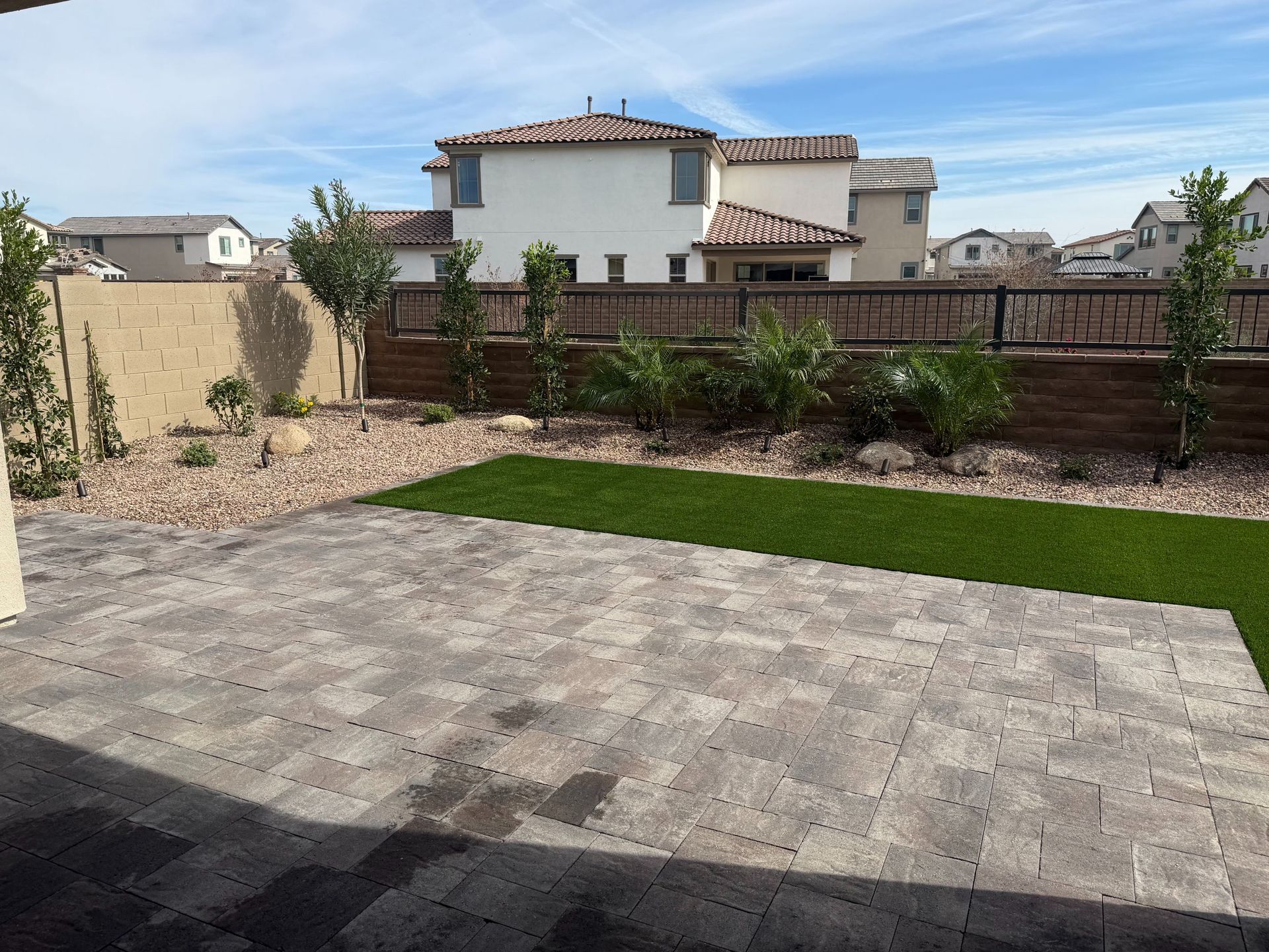 Backyard patio with stone pavers, lawn, shrubs, and a wooden fence behind a house