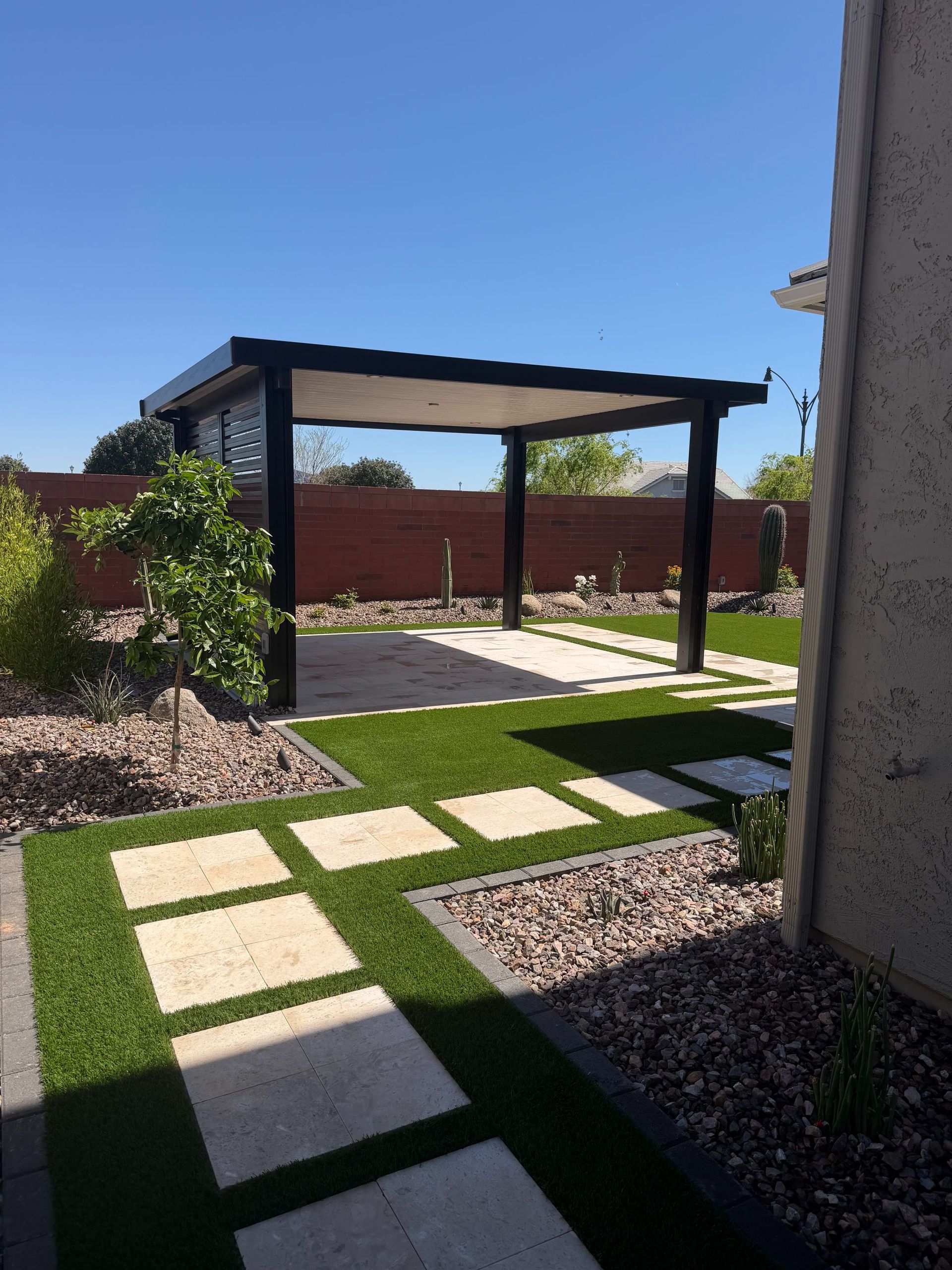 Modern backyard patio with pergola, stone path, green lawn, and red fence under a clear blue sky