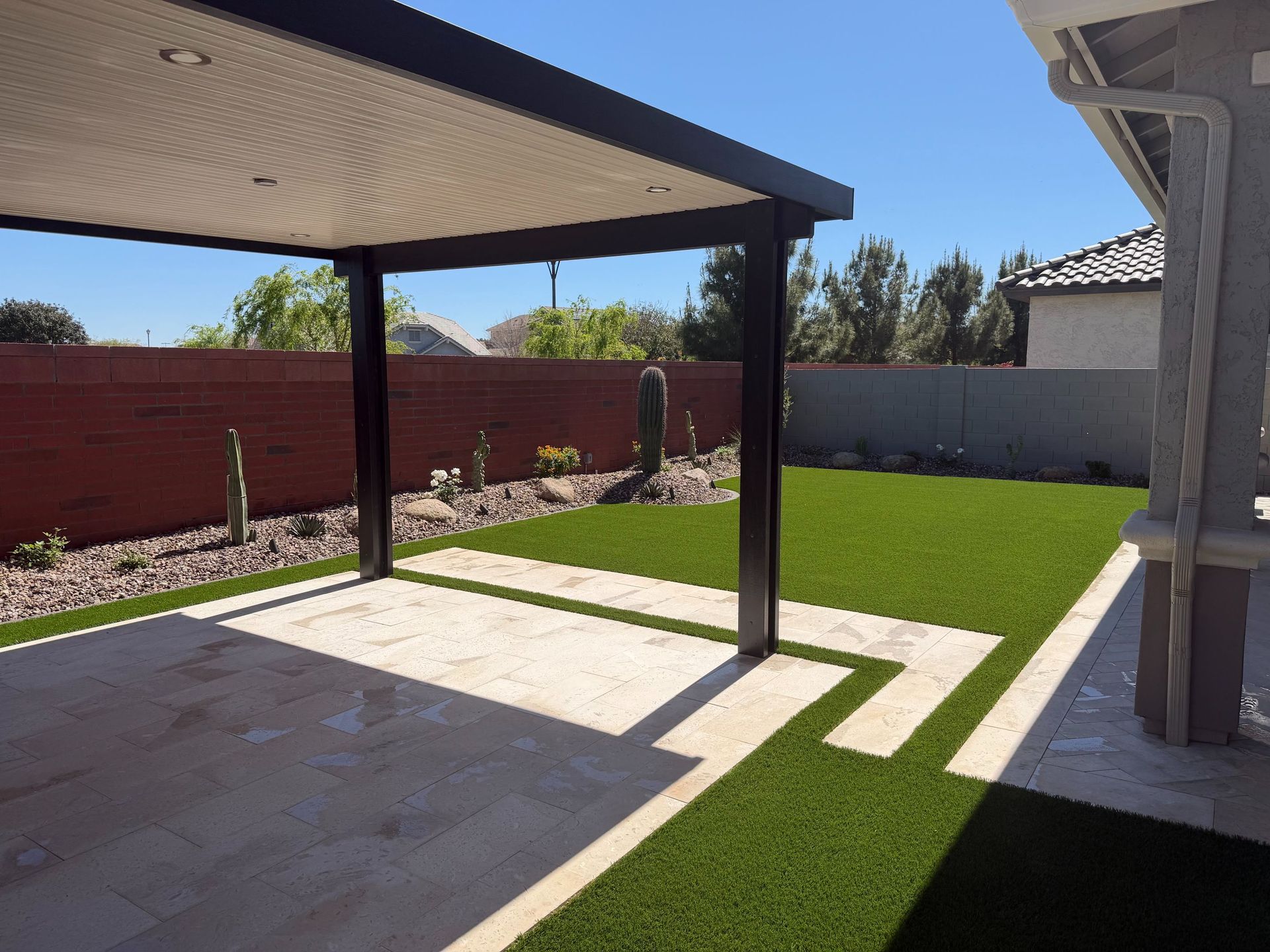 Backyard patio with a pergola, concrete walkway, green lawn, and red brick fence under a clear sky
