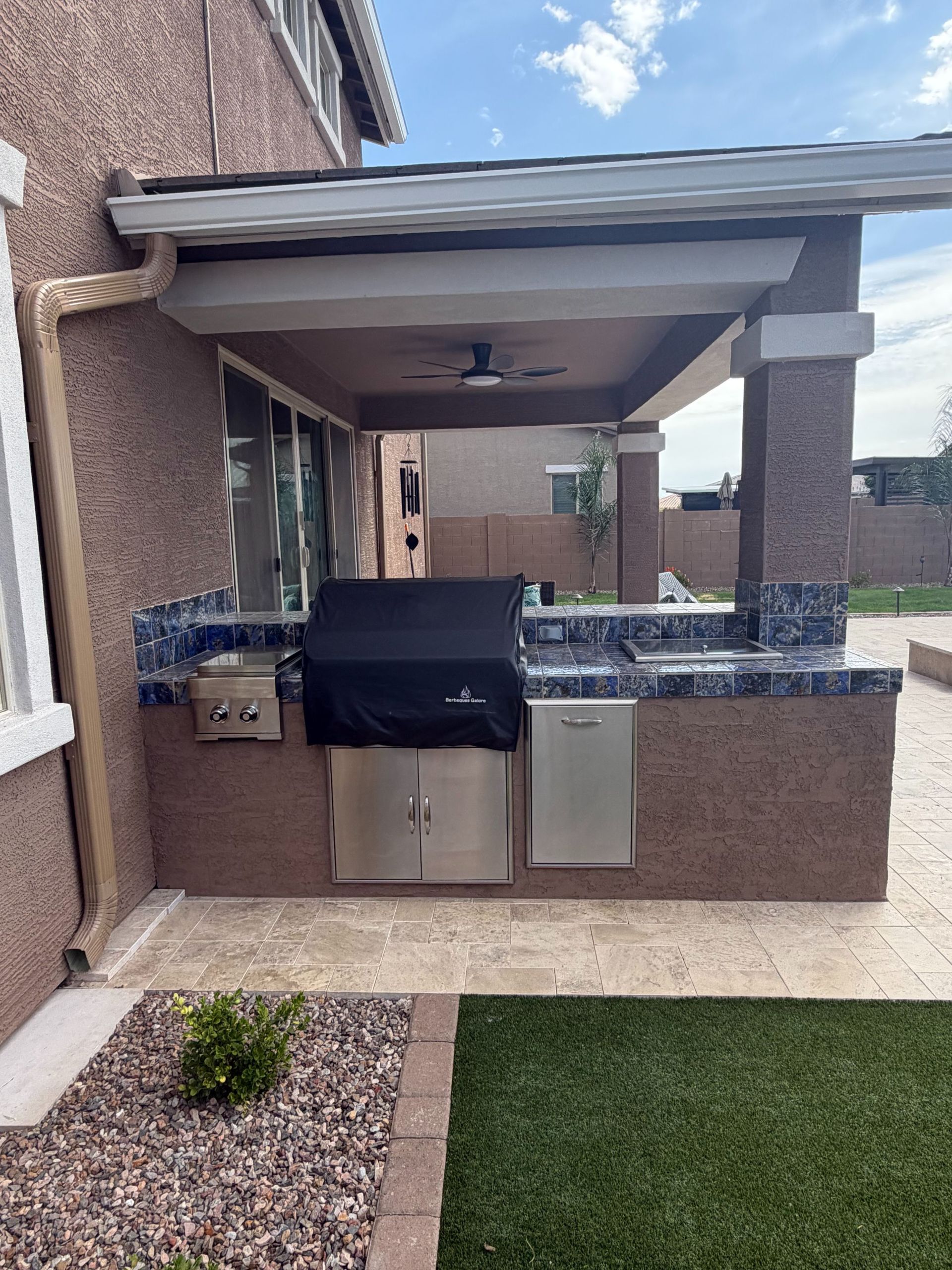 Covered outdoor kitchen with built-in grill, sink, and cabinets beside a patio.