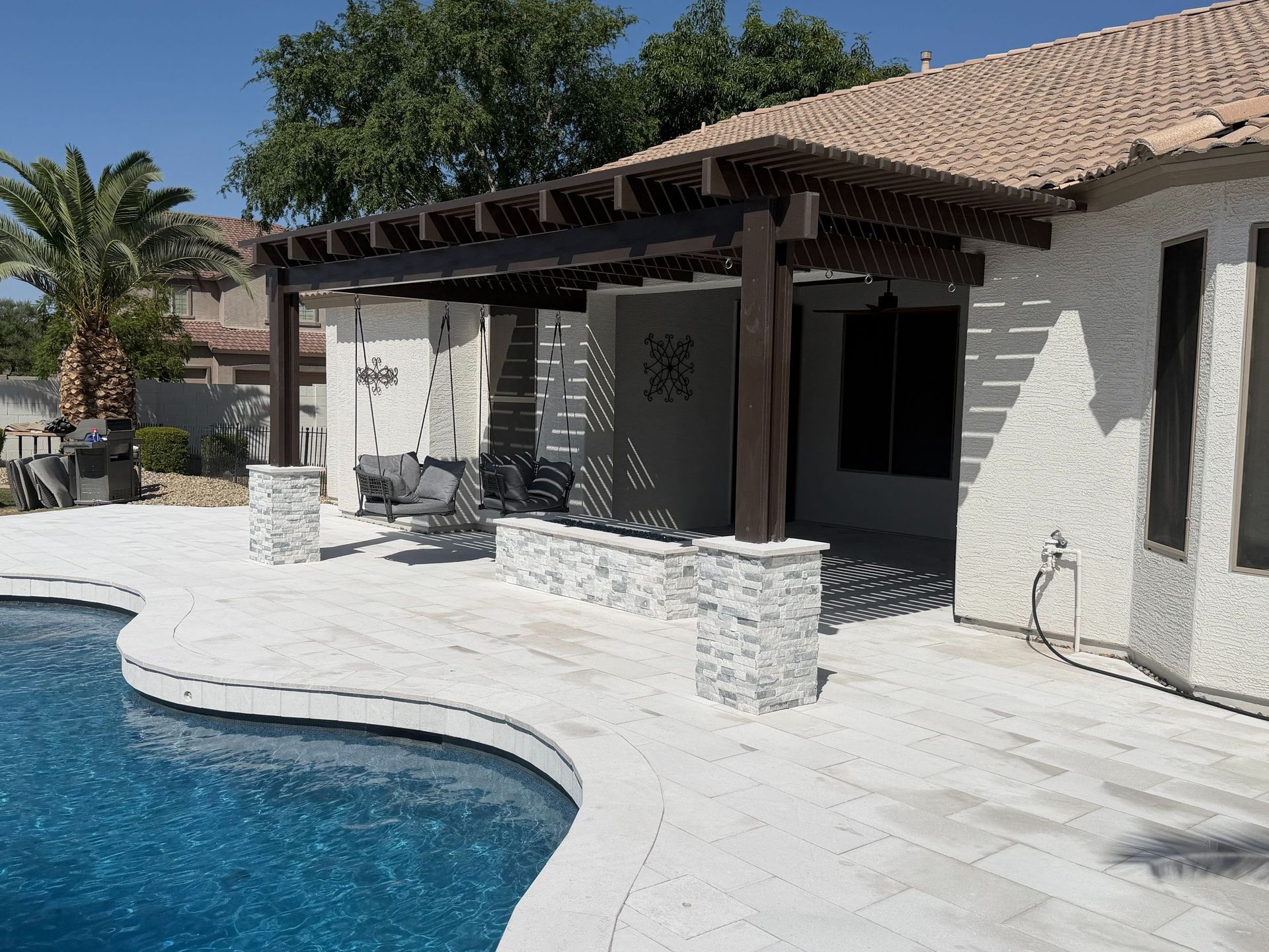 Poolside patio with white stone paving, covered terrace, and tan stucco house under a tiled roof.