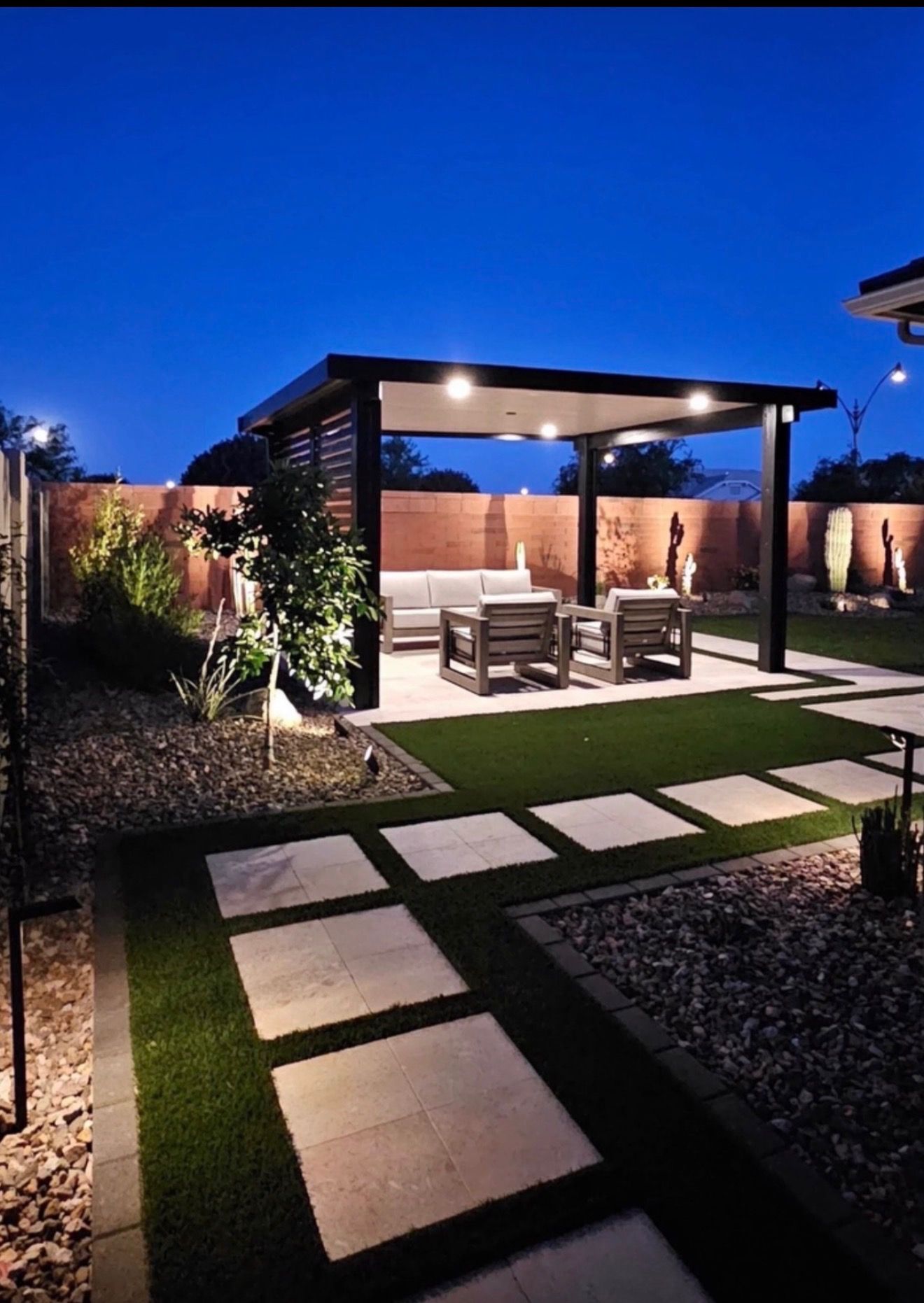 Modern backyard patio with pergola, lit seating area, and stepping-stone path at dusk