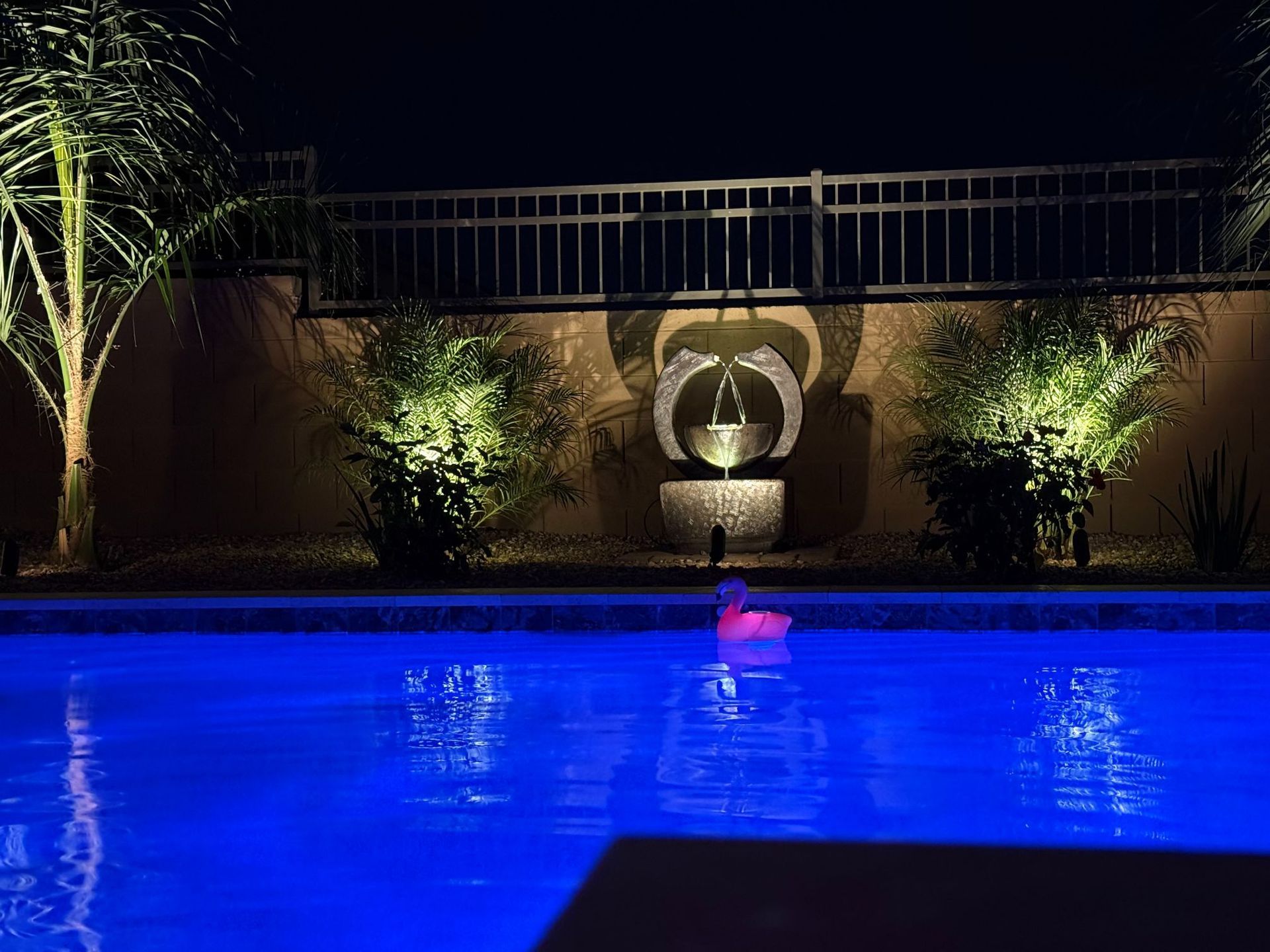 Night poolside scene with blue-lit water, illuminated plants, and a fountain against a dark wall