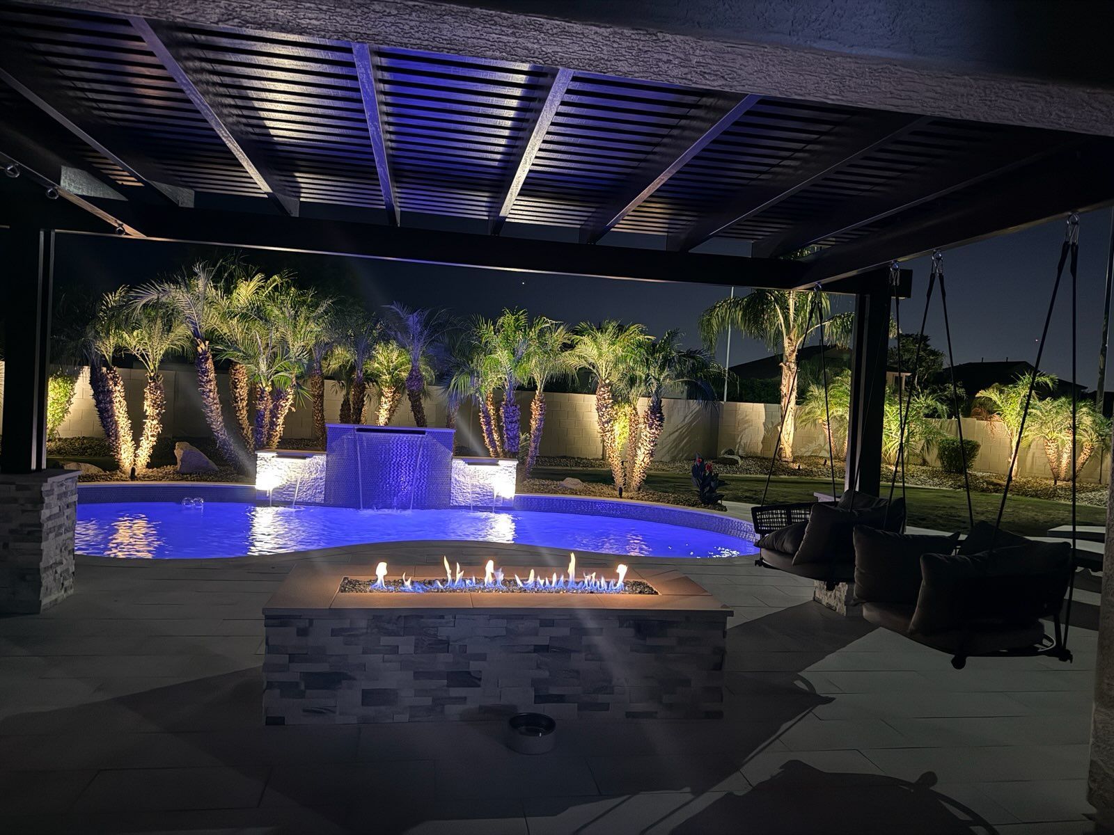Fire pit and lit pool under a covered patio at dusk, with palm trees and lounge chairs