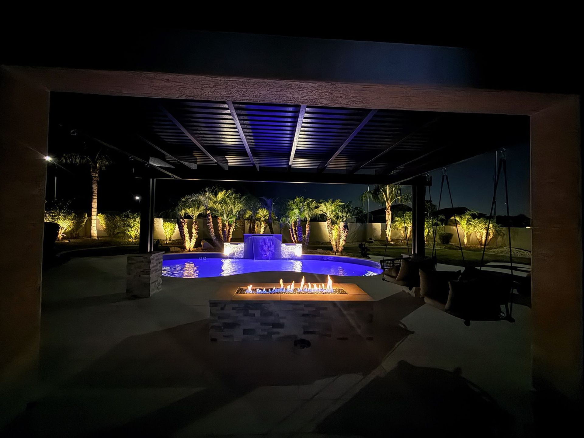 Nighttime patio with a lit fountain, blue accent lights, and a fire pit under a pergola