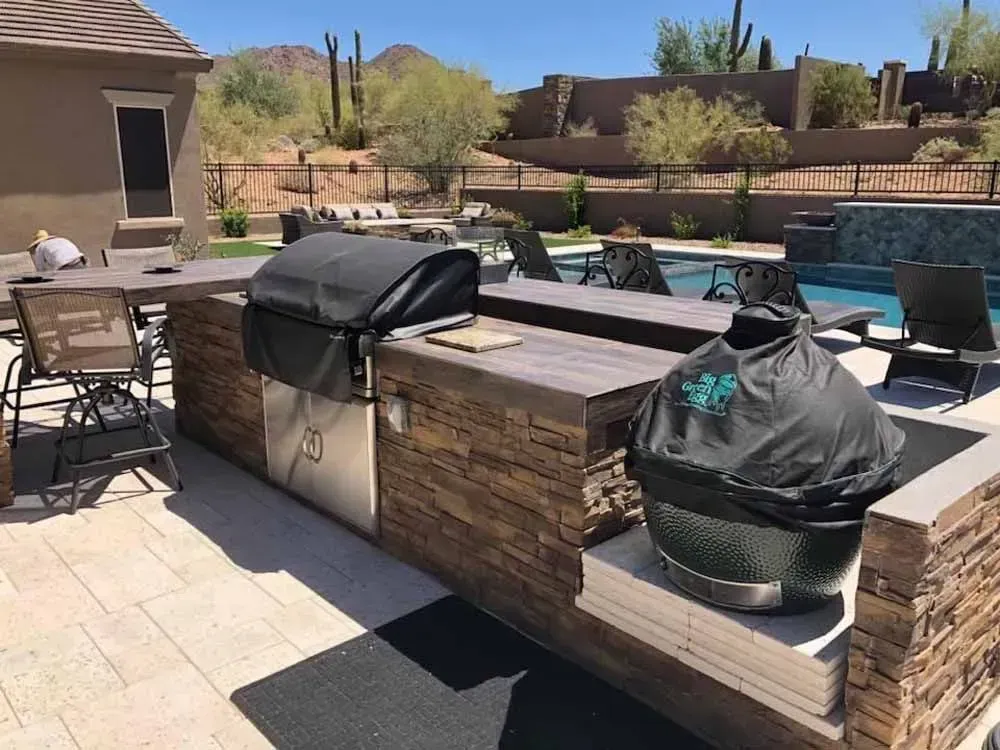 Outdoor kitchen with a grill, Big Green Egg, dining area, and pool in a desert setting.