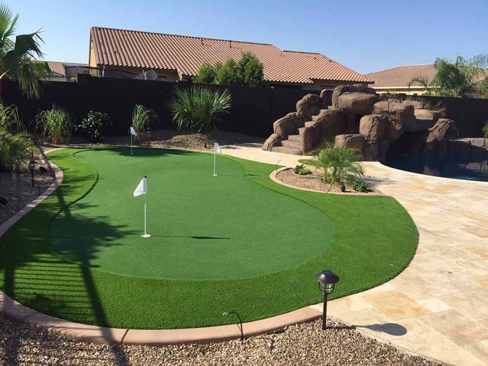 A backyard putting green with three flags, surrounded by artificial turf and landscaping.