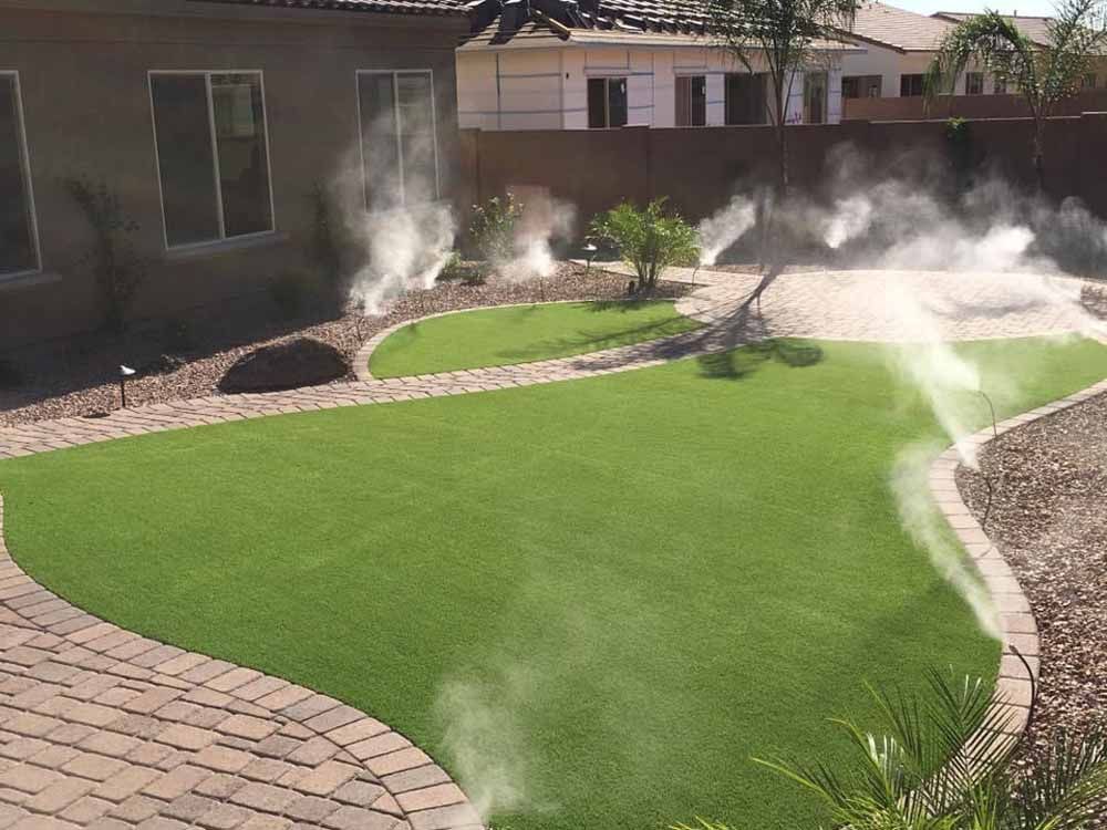 Lawn sprinklers misting an artificial green lawn in a backyard setting, with a paved walkway.