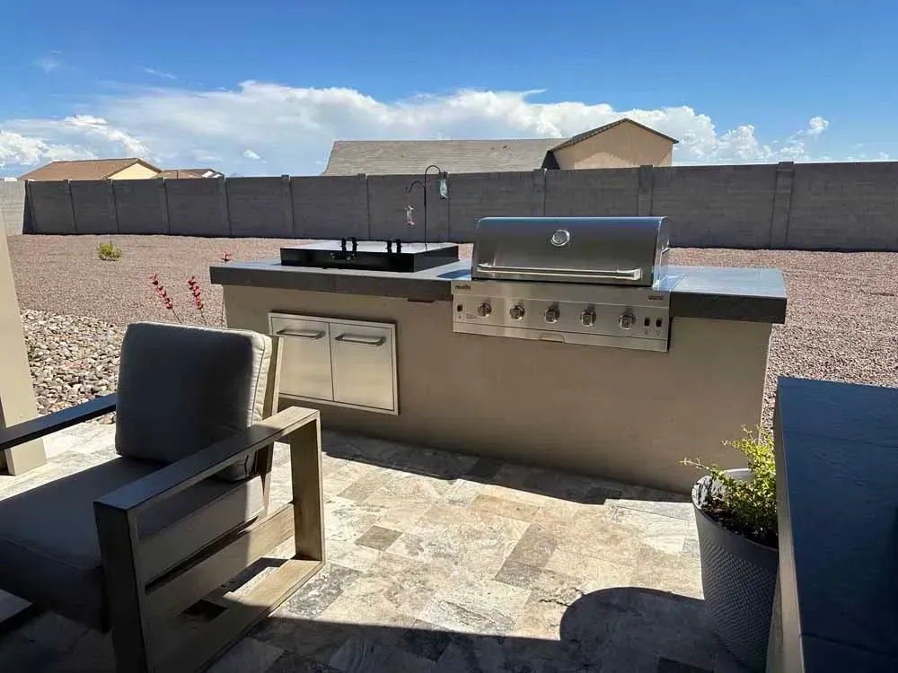 Outdoor kitchen with a grill, sink, and storage, on a tiled patio. A chair sits nearby.