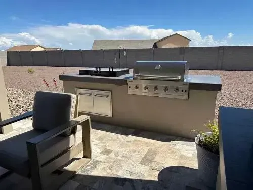 Outdoor kitchen with a grill, sink, and storage, on a stone patio with a chair.