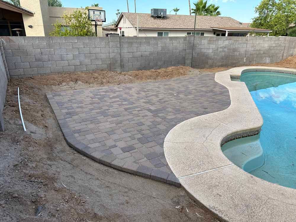 Backyard patio with brick pavers next to a swimming pool and a concrete block wall.