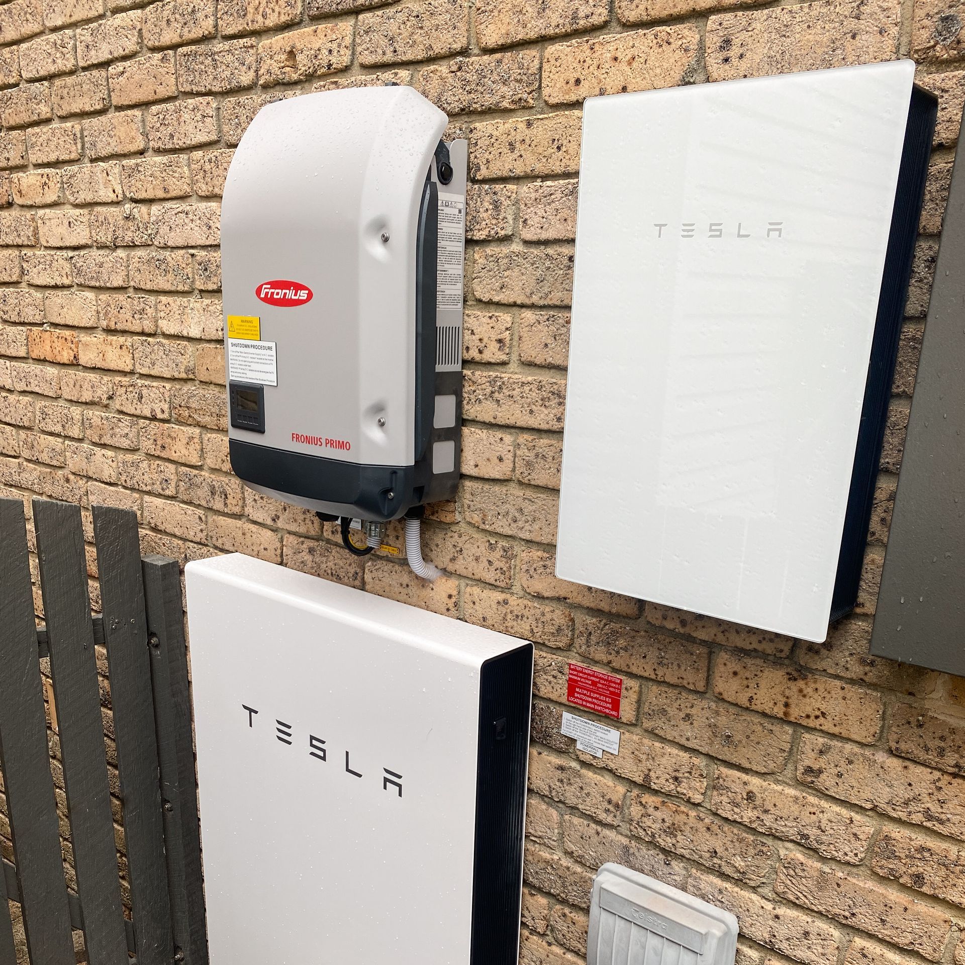 Tesla powerwall units mounted on an exterior brick wall beside a gray electrical meter box.