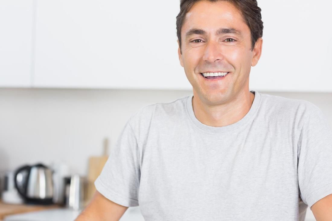 Man smiling in a kitchen wearing a light gray t-shirt. White cabinets and a kettle are in the background.