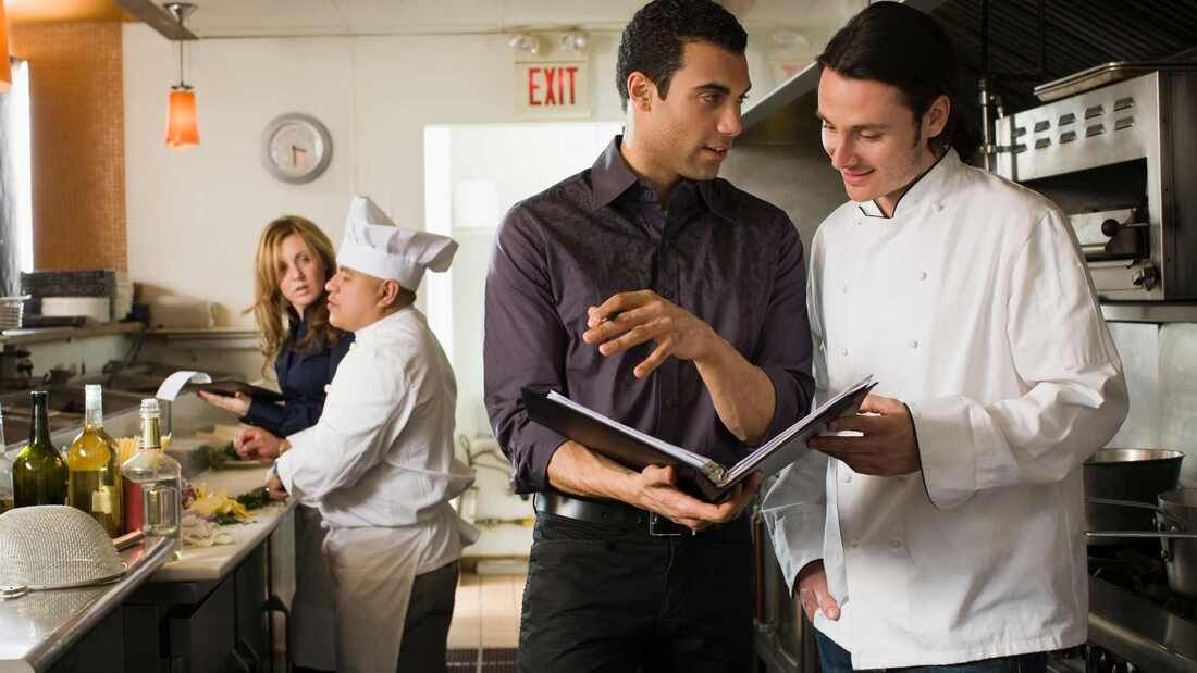 Restaurant kitchen: Two men reviewing a menu, chef in white coat, others preparing food.