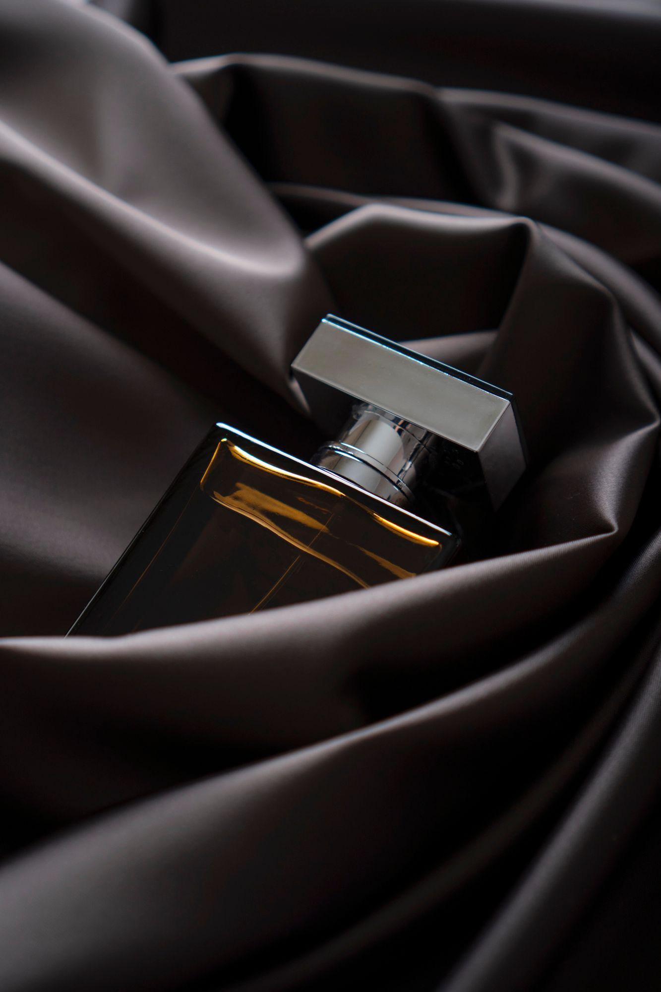 Frasco de perfume negro con tapón plateado, envuelto en pliegues de satén oscuro.