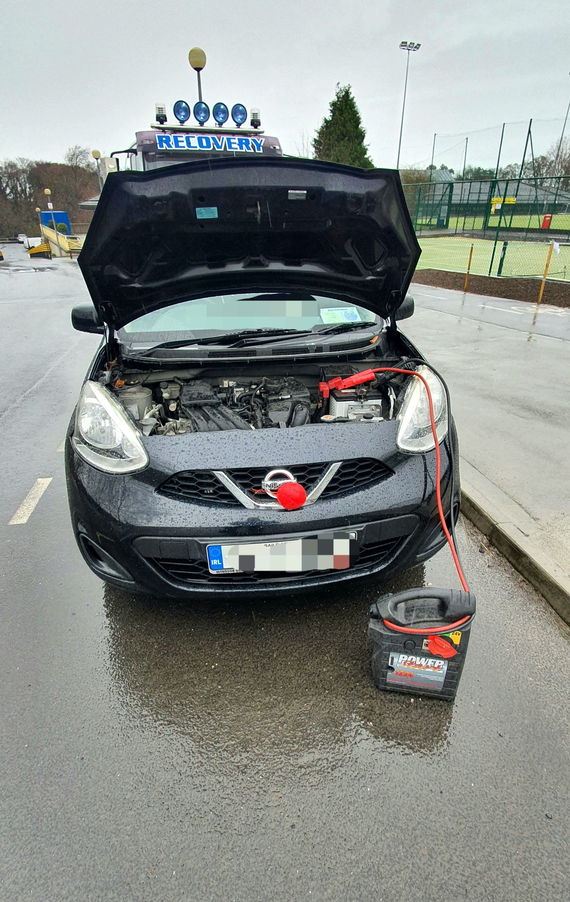 Black car with hood open on a wet road, battery charger attached, and a tool box on the ground.