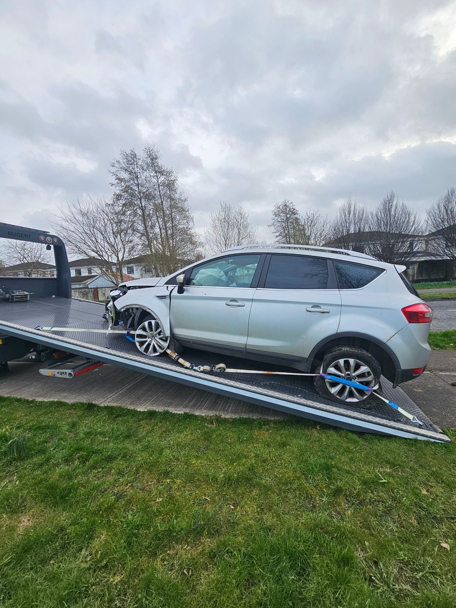 Silver SUV tipped on a tilted trailer ramp in a grassy yard under cloudy skies