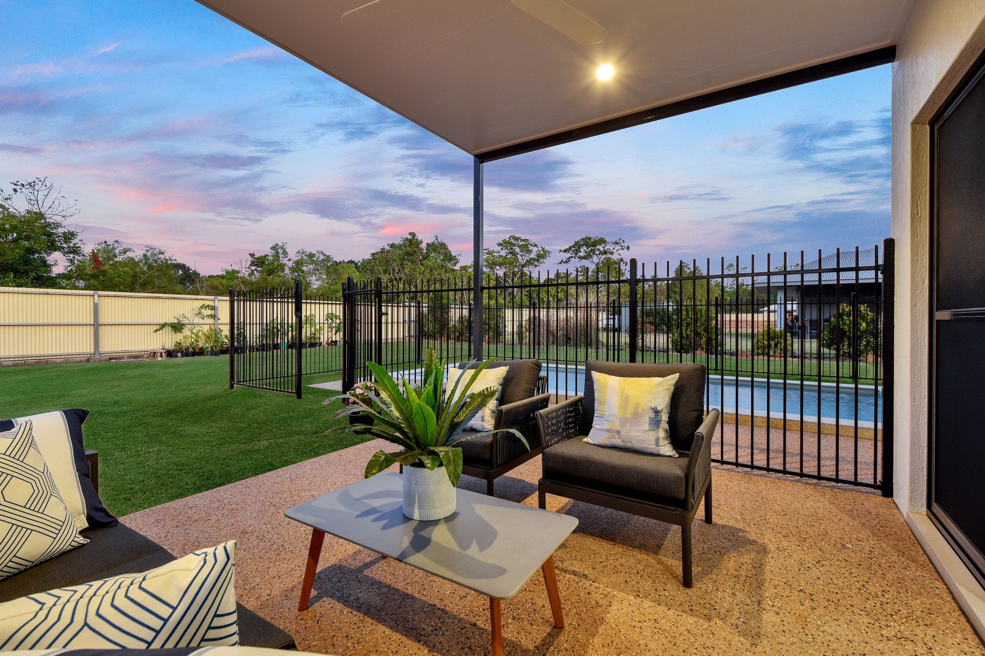 A patio with chairs , a table , and a fence with a pool in the background.
