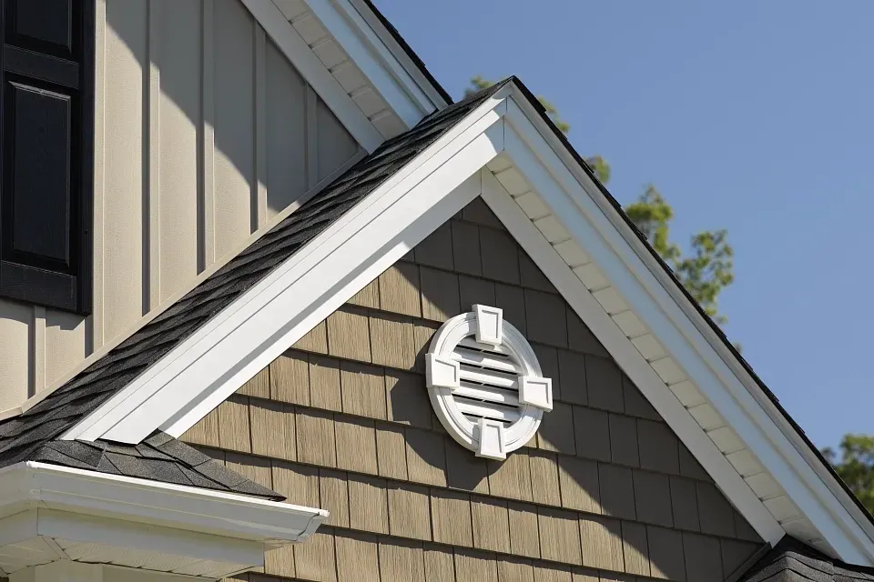 Close-up of a house gable with gray siding, white trim, and a round attic vent against a blue sky