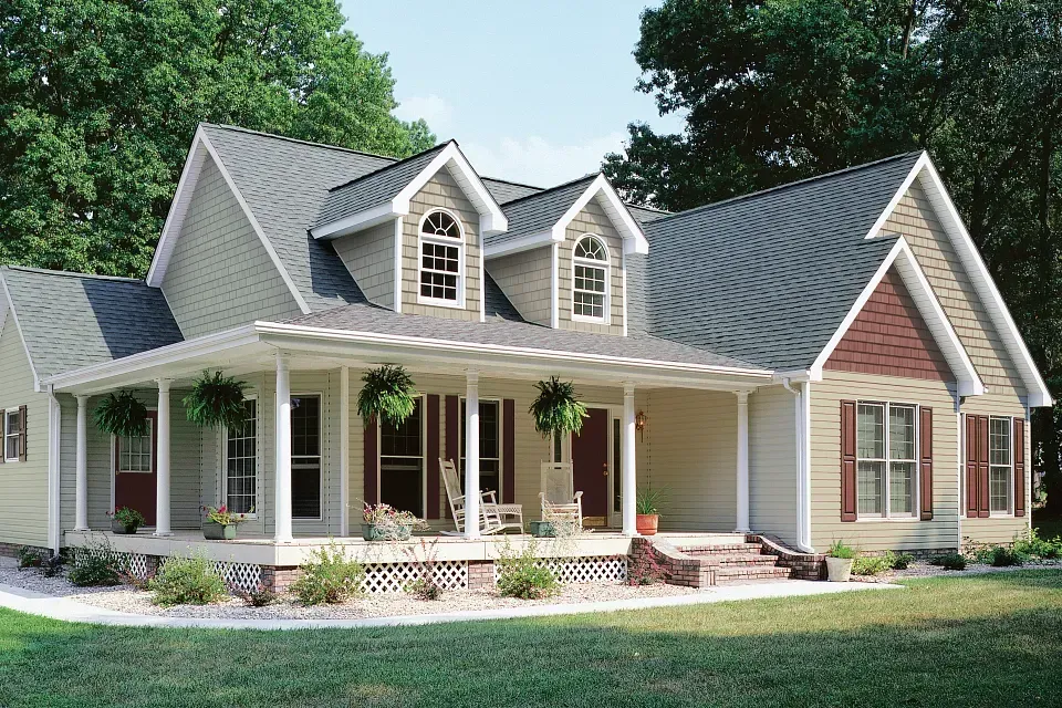 Charming beige house with a wraparound porch, gray roof, and white columns surrounded by green trees.