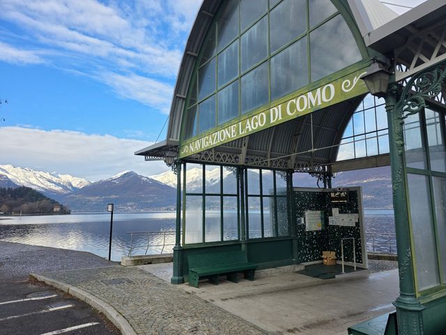 Una stazione degli autobus verde con un lago e montagne innevate sullo sfondo, visibile la scritta "LAGO DI COMO".