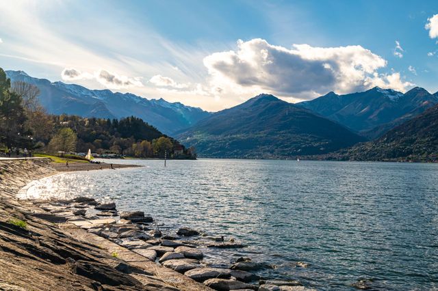 Lago circondato da montagne sotto un cielo azzurro parzialmente nuvoloso. Costa rocciosa in primo piano.