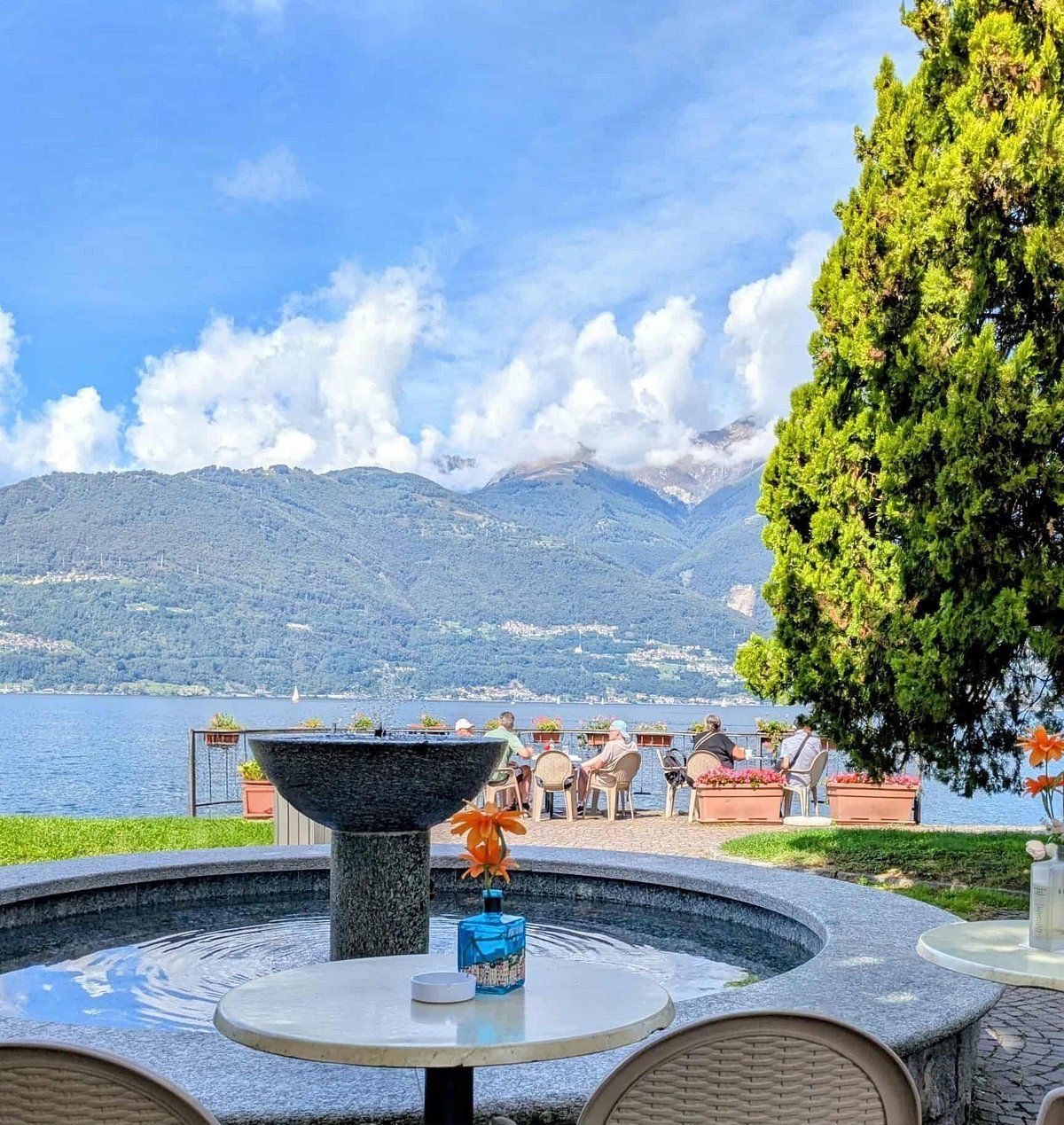 Un bar in riva al lago con una fontana in primo piano e montagne sullo sfondo sotto un cielo azzurro.