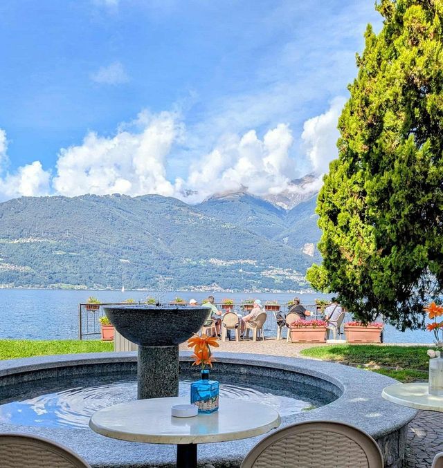 Un bar in riva al lago con una fontana in primo piano e montagne sullo sfondo sotto un cielo azzurro.