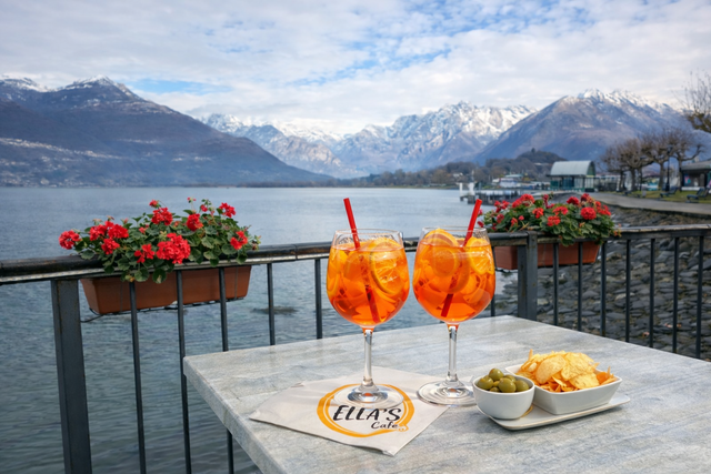 Due Aperol Spritz su un tavolo con un lago e montagne sullo sfondo. Si vedono anche olive e patatine fritte.