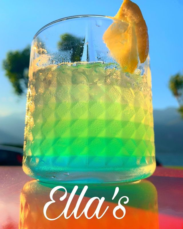 Cocktail a strati arcobaleno in un bicchiere con guarnizione di limone; sulla base c'è scritto Ella's.