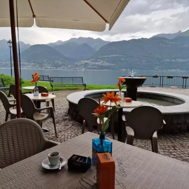 Bar all'aperto con vista sul lago e sulle montagne, tavoli con bevande e fiori, cielo nuvoloso.