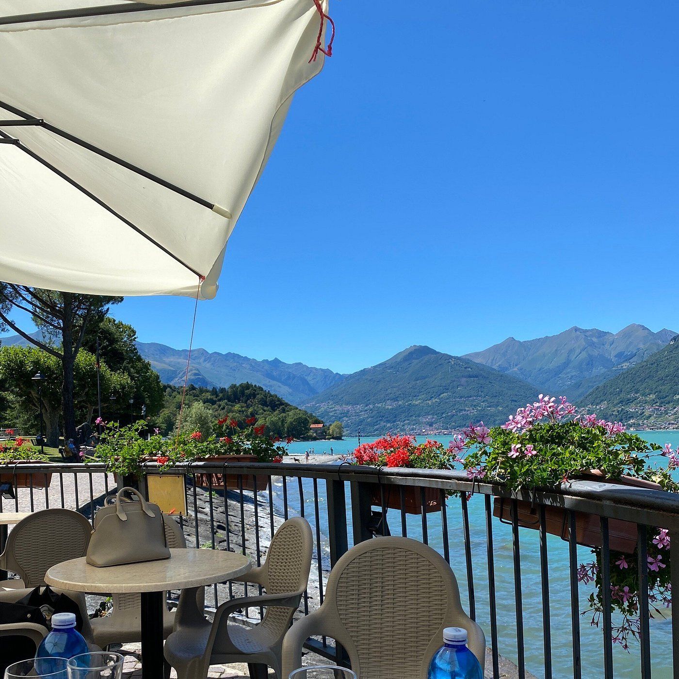 Bar all'aperto con vista sul lago e sulle montagne sotto un cielo azzurro; tavolo, sedie, fiori e ombrellone visibili.