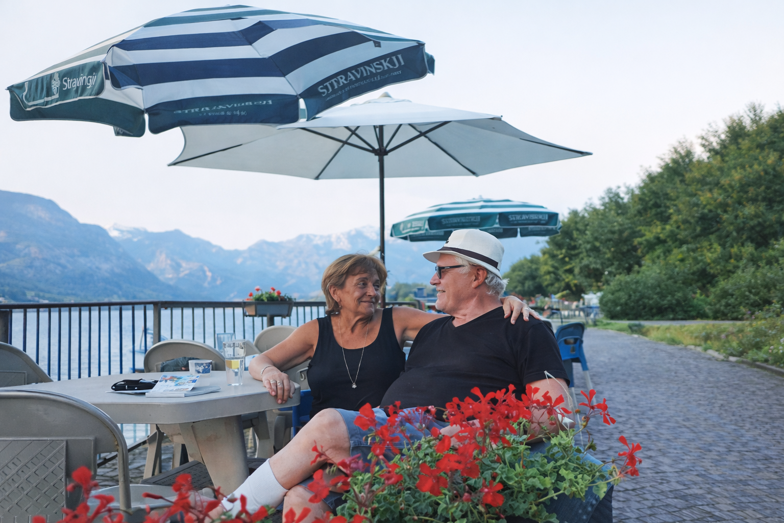 Coppia seduta in un bar all'aperto con vista sul lago e sulle montagne. Sono sotto gli ombrelloni, vicino a fiori rossi.