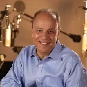 Man smiling in a blue shirt, seated in a recording studio with microphones.