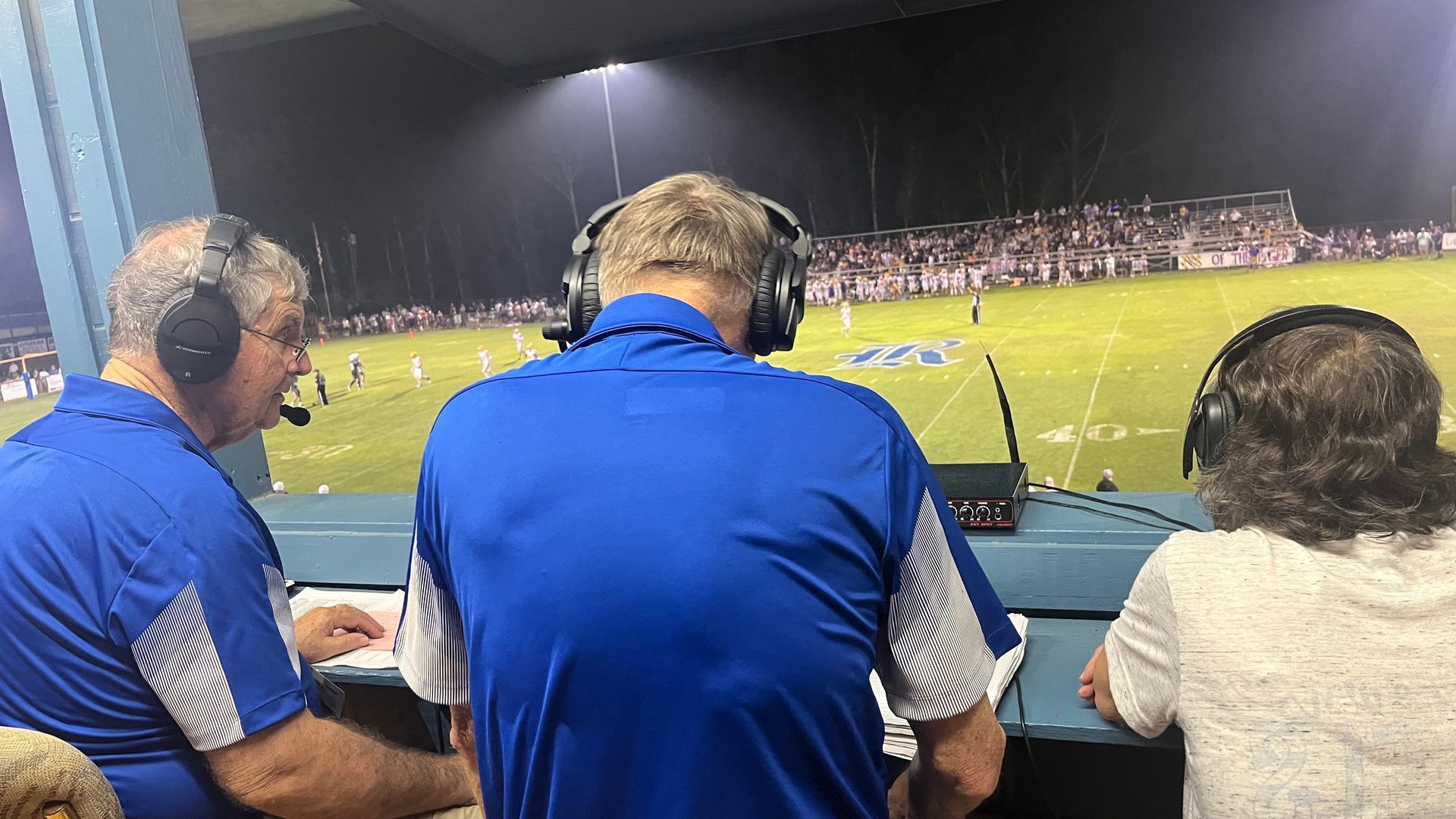 Three sports commentators in blue shirts watching a lit soccer field from a press box