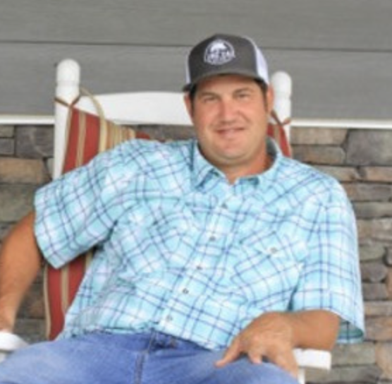 Man in a plaid shirt and cap sits in a rocking chair on a porch, smiling.