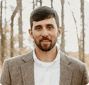 Man with dark hair and beard, wearing a brown blazer and white shirt, smiling outdoors.