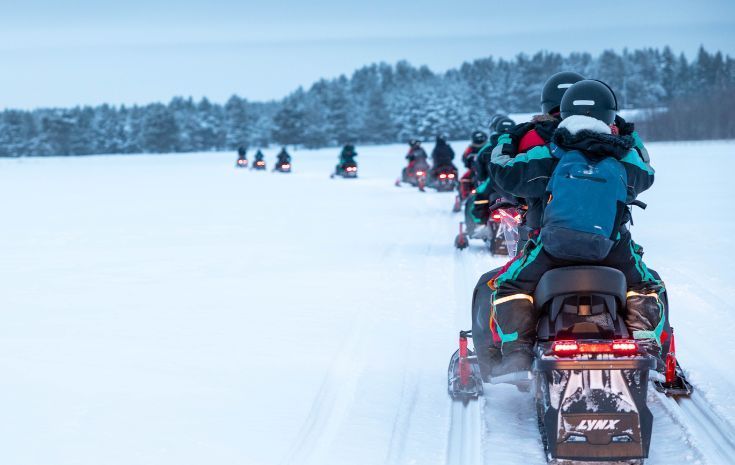Snowmobiles riding in a line across a snowy field near a forest