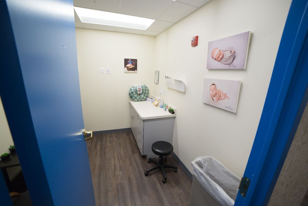 A doctor's exam room with a desk, stool, and baby-themed artwork on the walls. The door is blue.