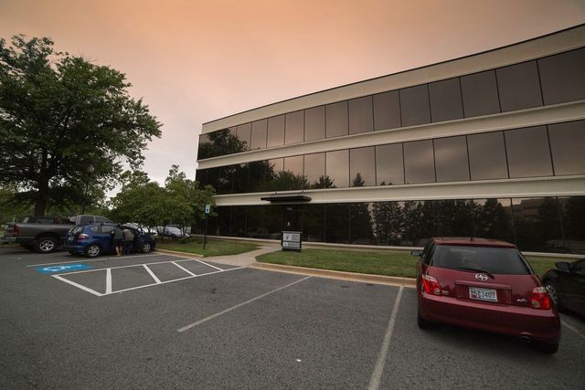Office building with a glass facade and a parking lot. Red car parked on the right, blue truck in the left, and a wheelchair parking spot.