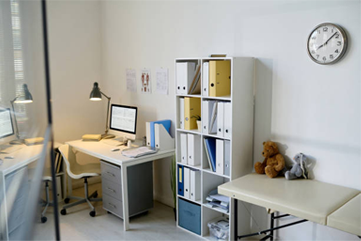 Doctor's office: desk with computer, files, clock on wall, examination table, and teddy bear.