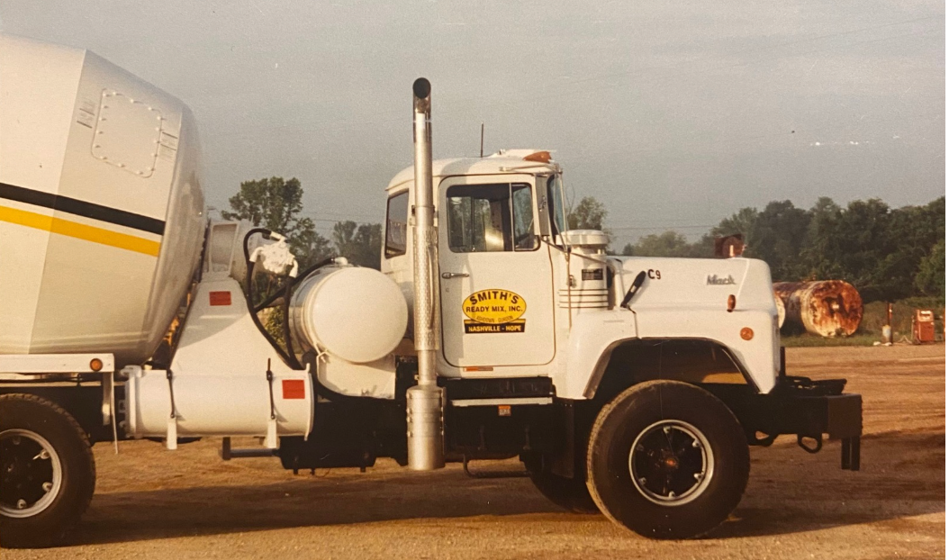 White Mack concrete mixer truck on a dirt lot. The truck has a black stripe and the company logo on the door.